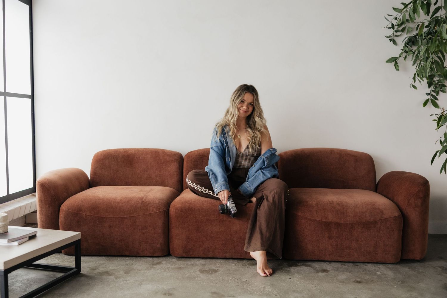 Woman in denim jacket and wide-leg pants sitting on a rust-colored modular sofa in a modern minimalist living room.