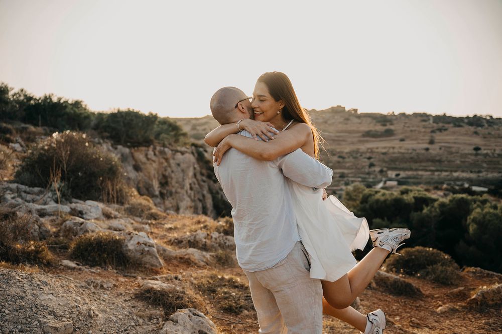 Couple embracing and laughing at sunset on a rocky hillside, man lifting woman in white dress outdoors.