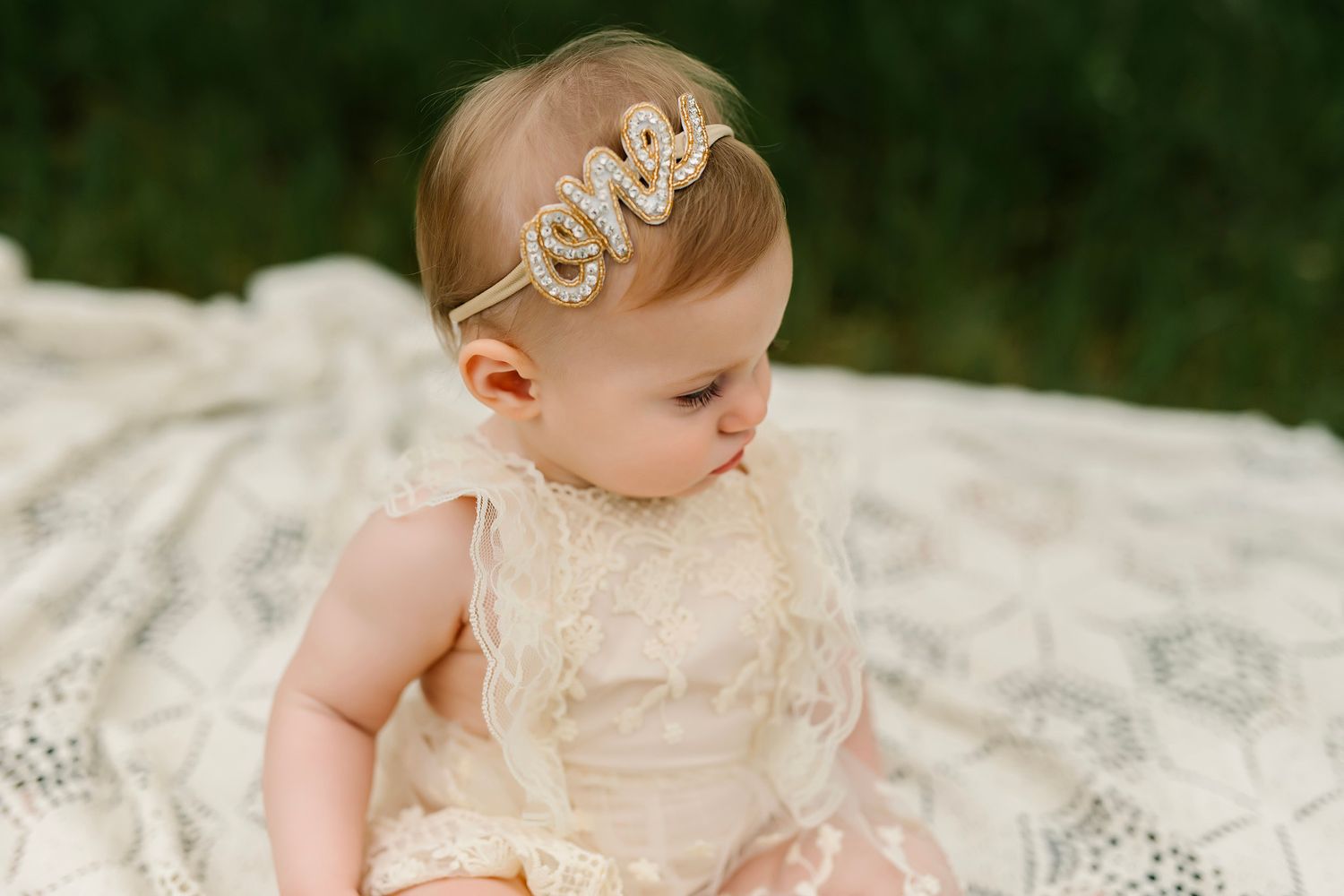 Baby girl in lace dress and gold 'one' headband sits on white blanket for first birthday photo.