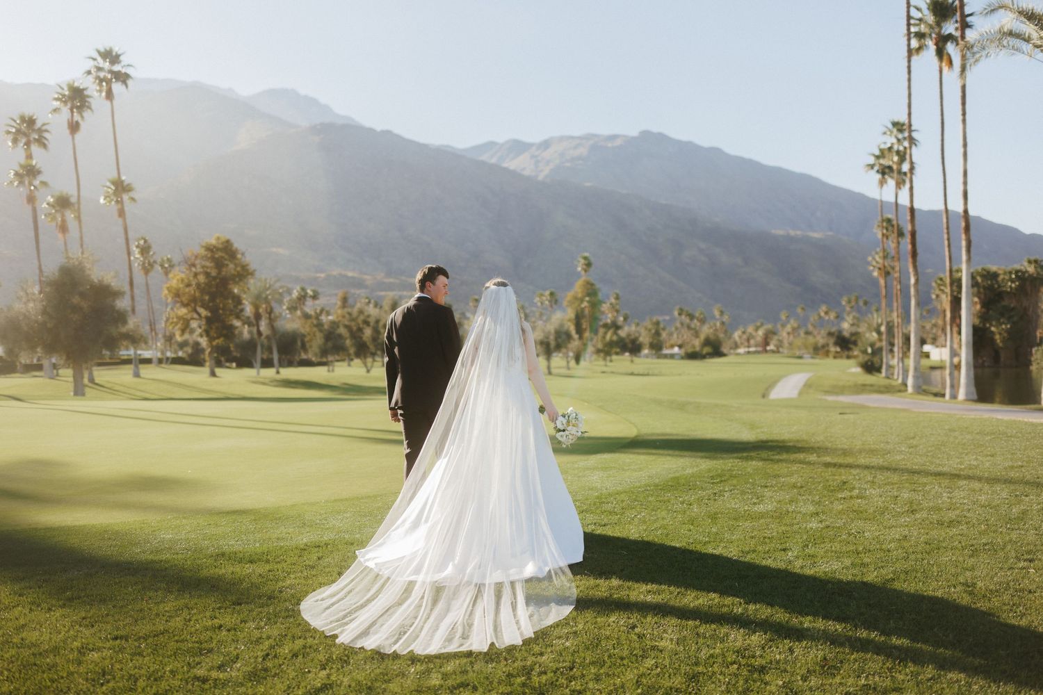 Bride and groom stand on lush green golf course with stunning mountain backdrop and palm trees in Palm Springs.