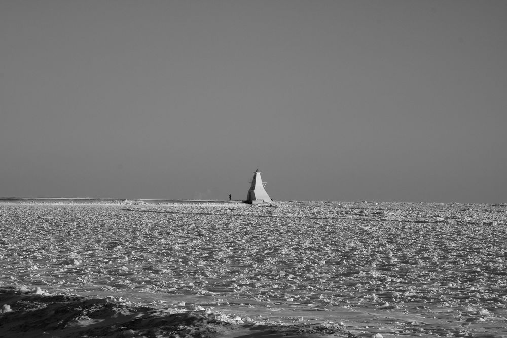 A black and white panoramic seascape shows a distant lighthouse on the horizon with sparkling water in the foreground.