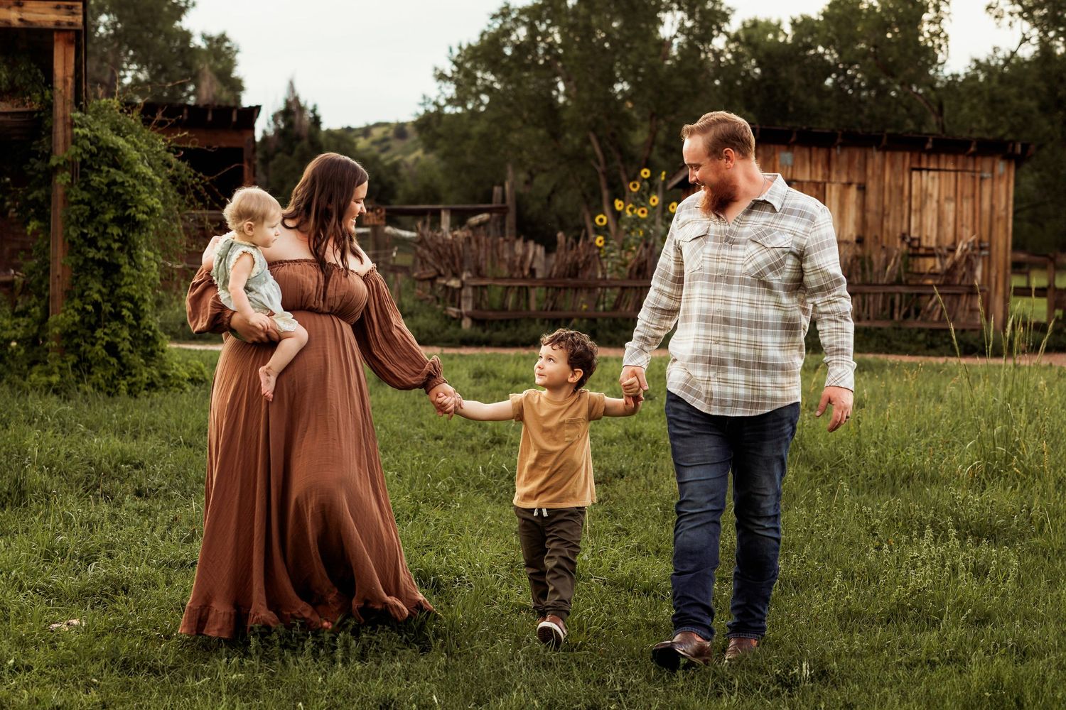 A family holds hands while walking together in a grassy backyard at sunset near rustic wooden buildings.