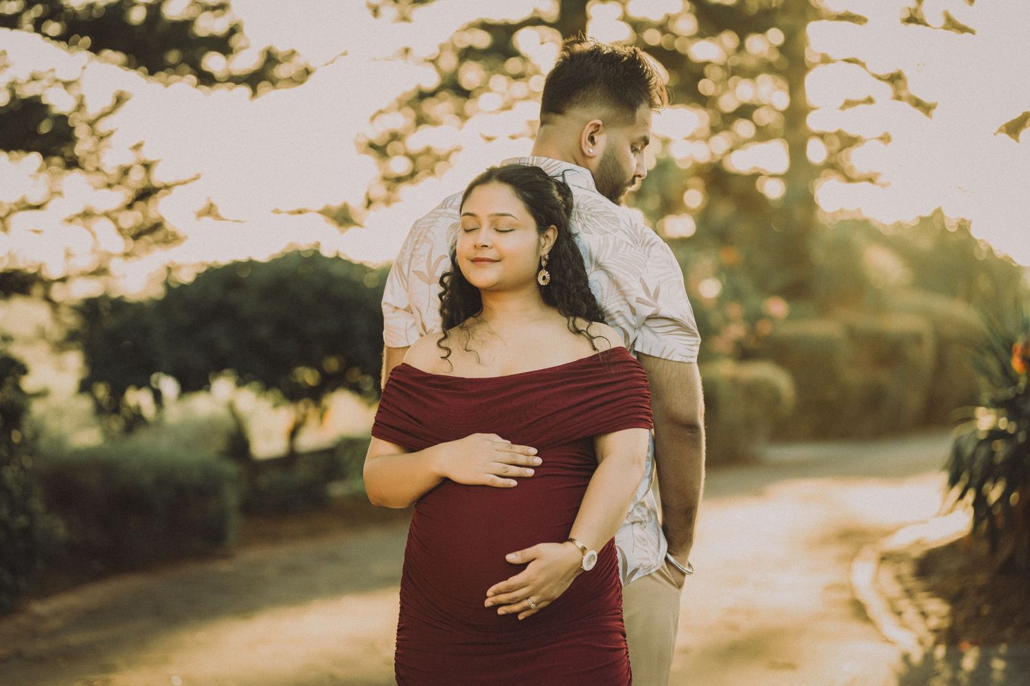 Couple sharing a tender maternity moment outdoors at golden hour, woman in red dress cradling baby bump.