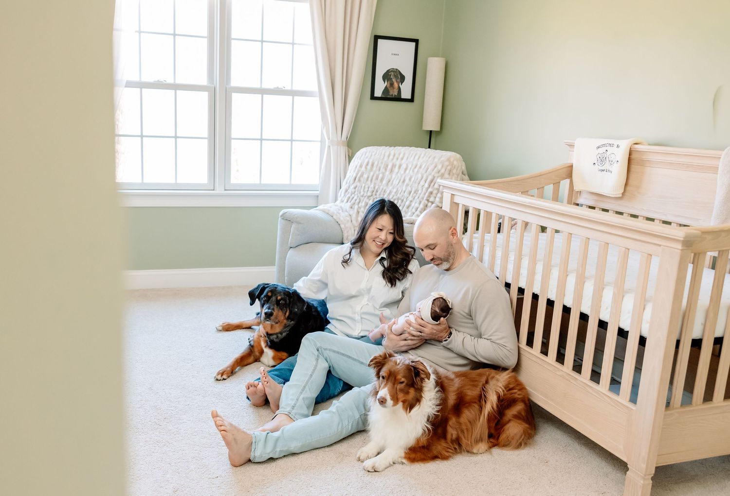 Family with newborn baby sitting on nursery floor with two dogs beside a white crib.