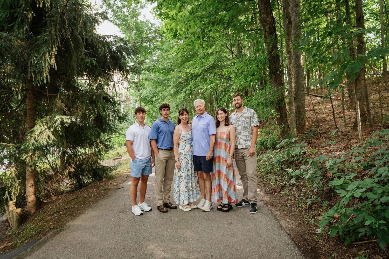 Family of six posing together on a tree-lined path surrounded by lush green trees on a sunny day.