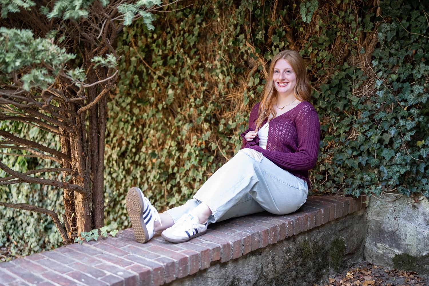 A person in a purple sweater sits on a stone wall surrounded by lush green foliage in an outdoor setting.