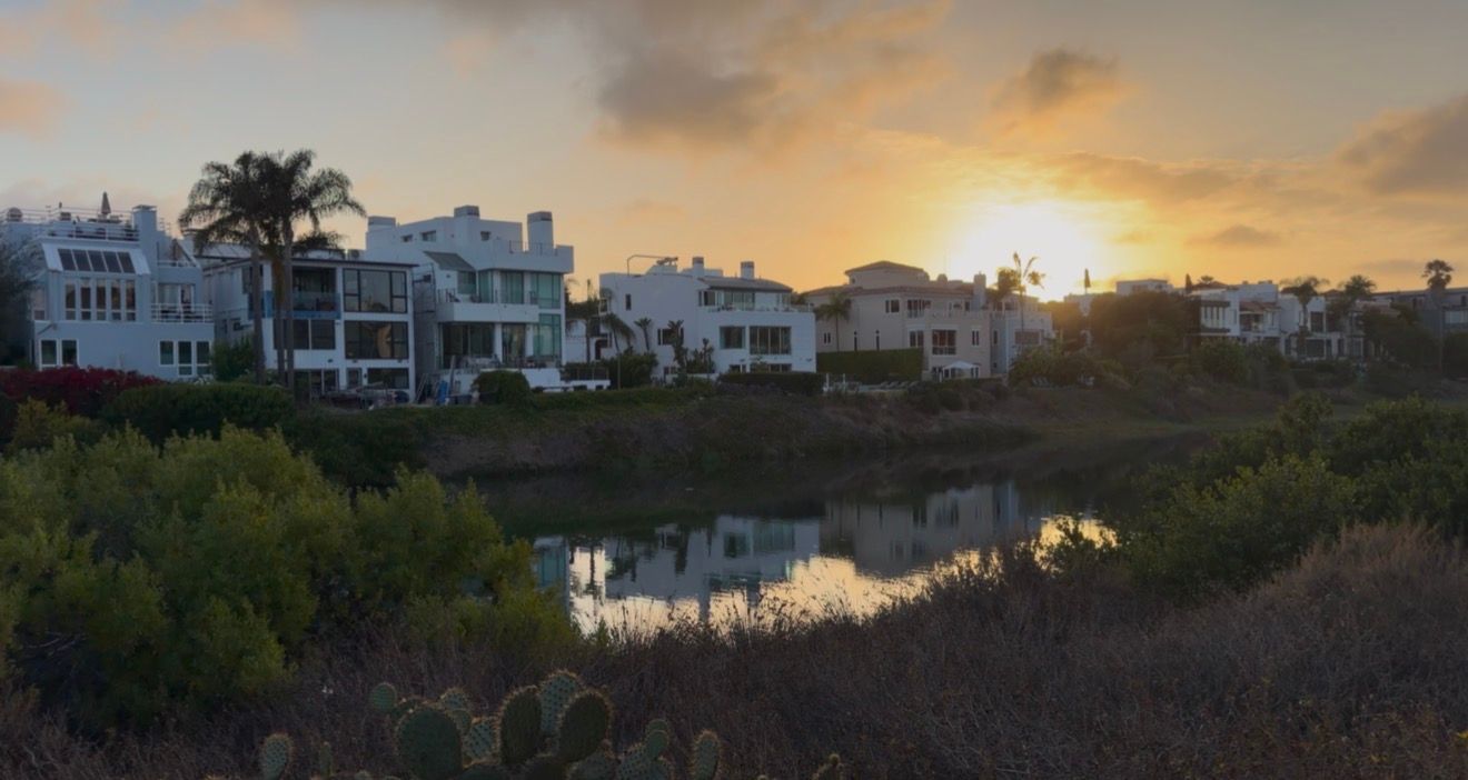Sunset over coastal homes reflecting in a calm lagoon surrounded by lush greenery and cacti.