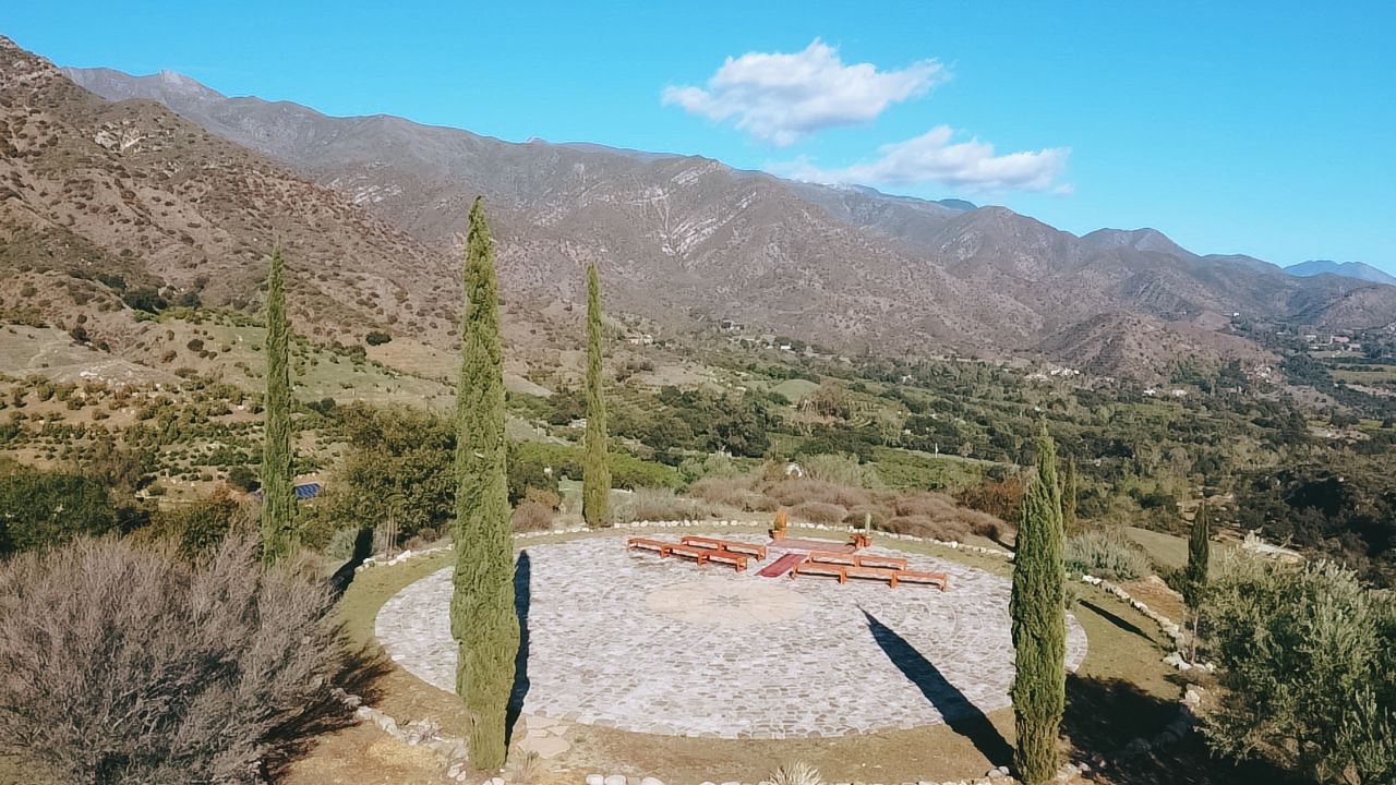 Aerial view of a circular stone patio with red benches and cypress trees overlooking a vast mountain valley landscape.
