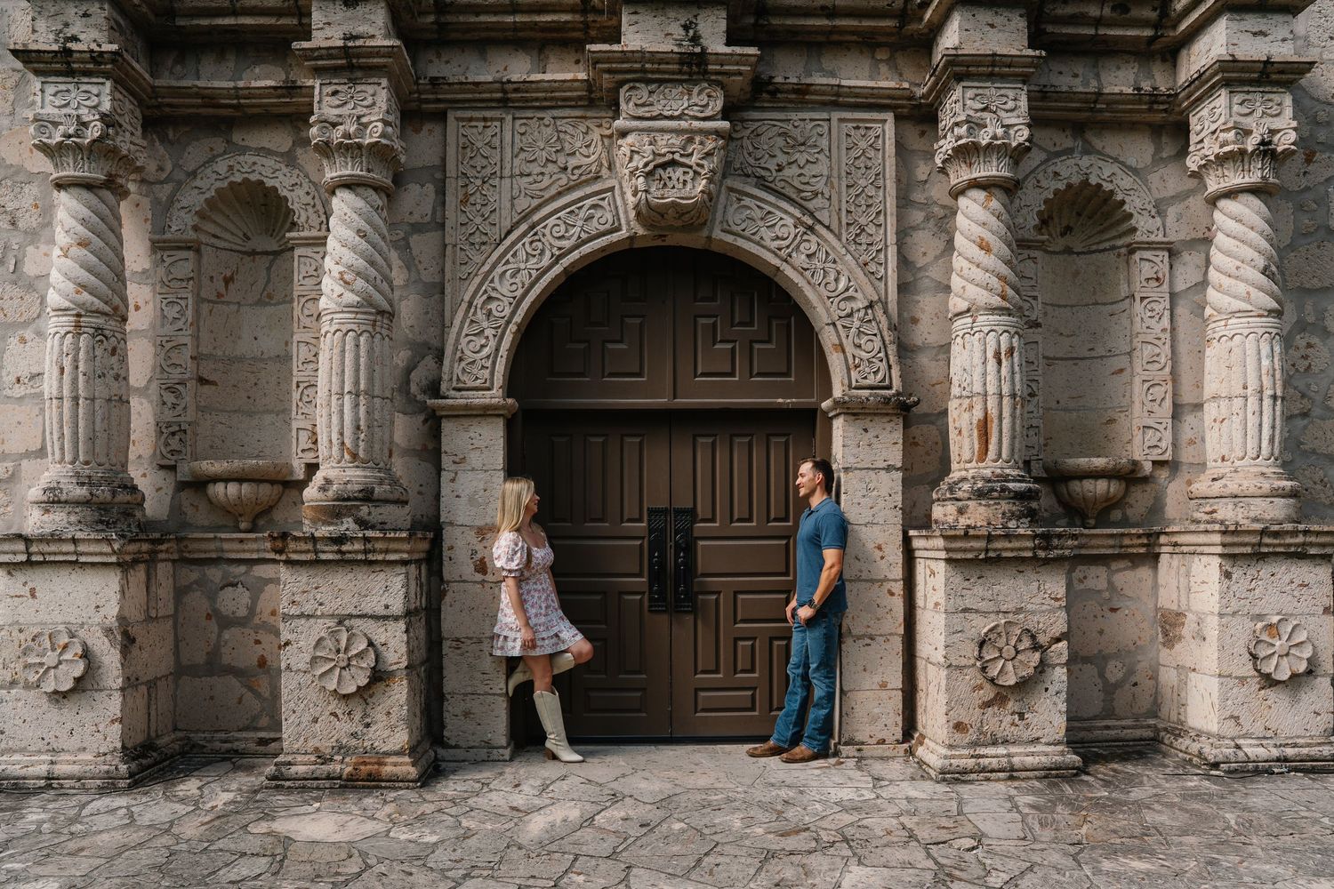Couple standing apart at ornate carved stone church doorway during outdoor engagement photo session.
