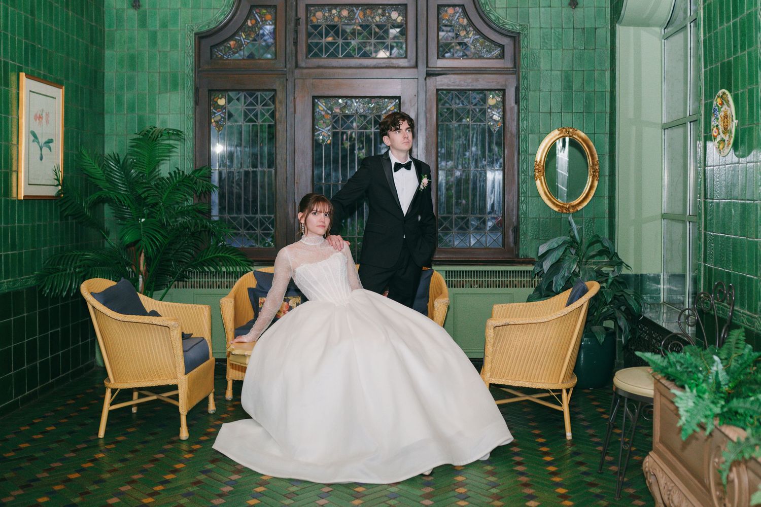 Bride in white ball gown sits beside groom in black tuxedo in elegant green-tiled room with yellow chairs.