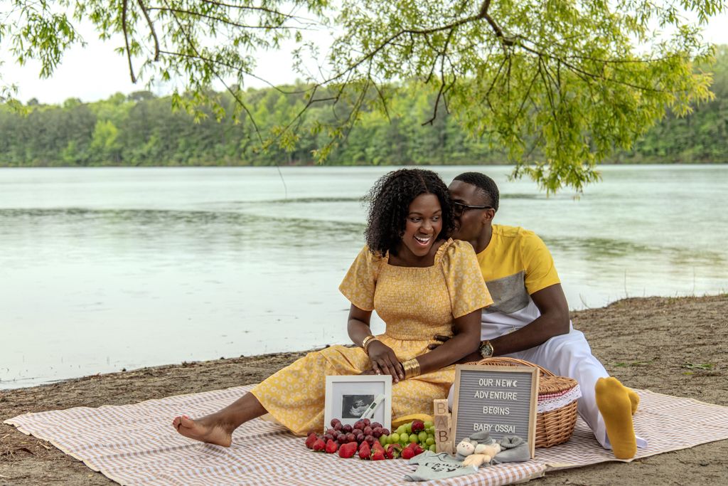 Couple in yellow outfits enjoying a romantic lakeside picnic with fruit, photos, and a letter board sign.