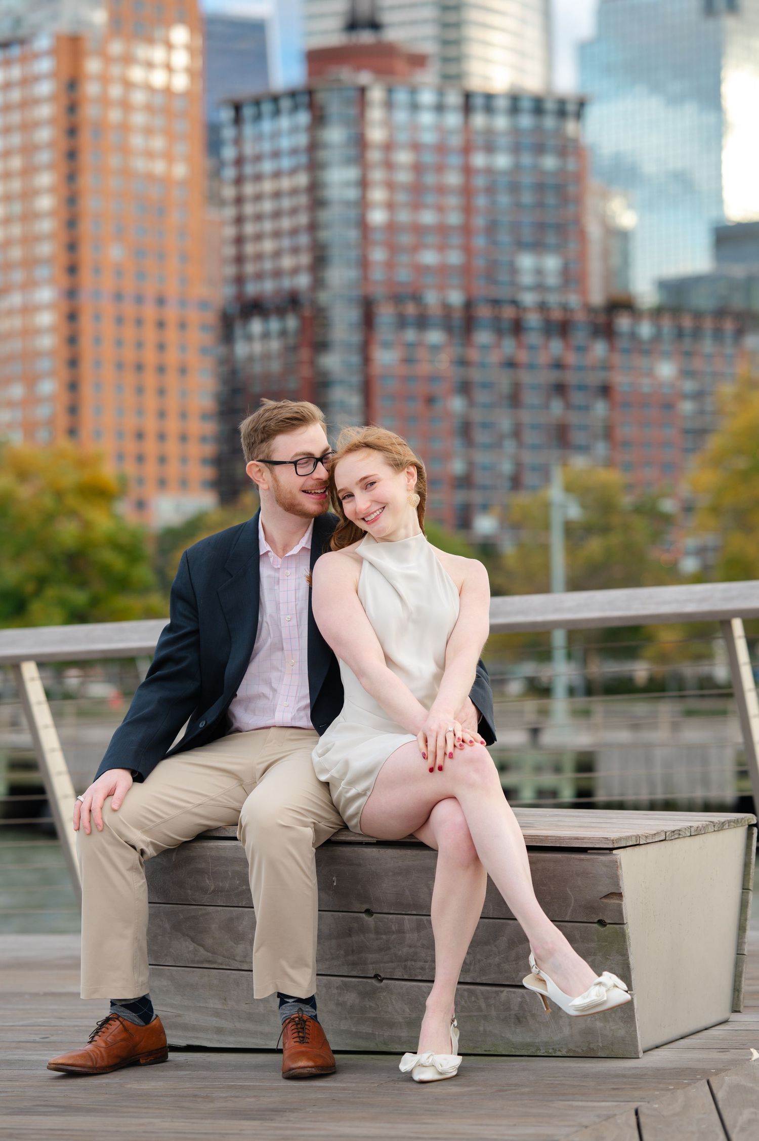 Couple sitting on wooden bench with city skyline backdrop, woman in white dress, man in navy blazer.
