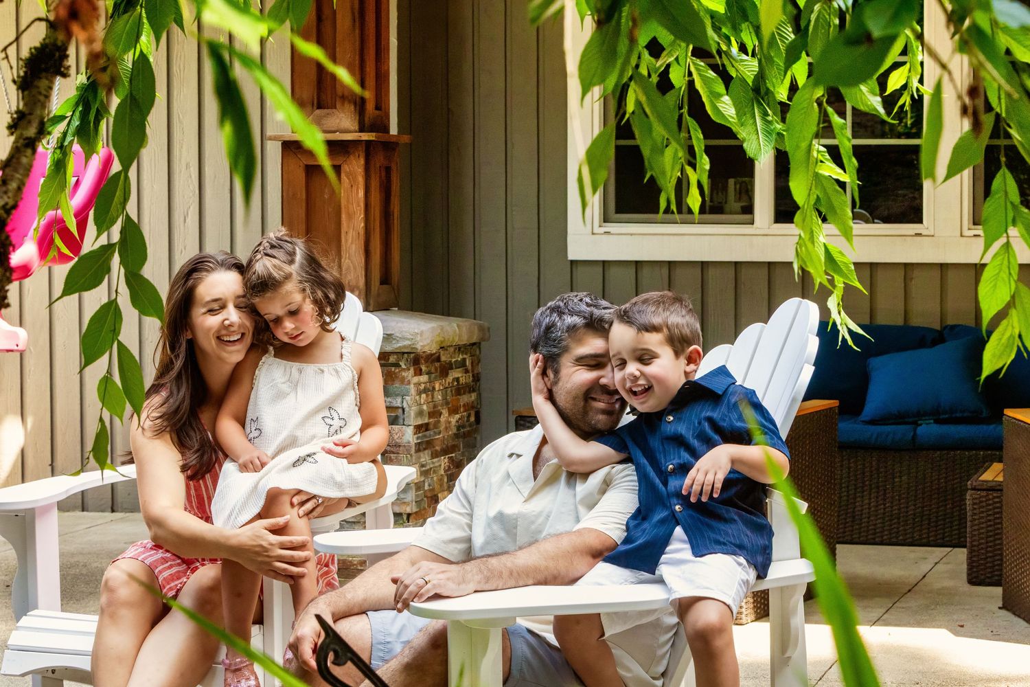 Happy family of four relaxing on a white porch chair outdoors, surrounded by lush green foliage on a sunny day.