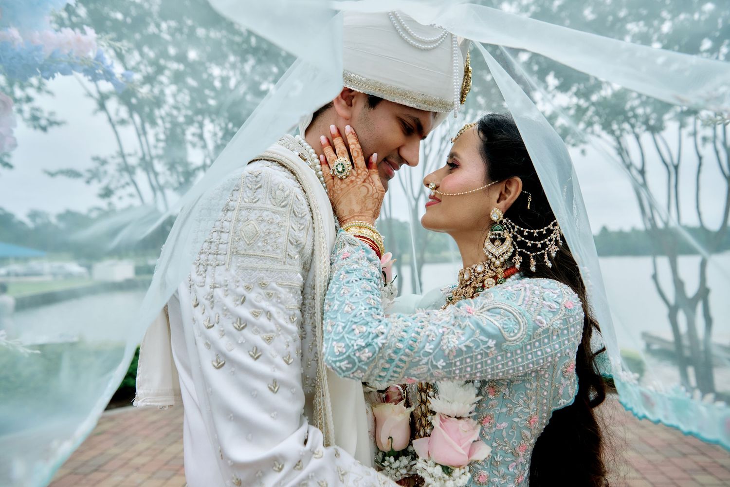 South Asian bride and groom in traditional white wedding attire share a romantic moment under a sheer canopy.