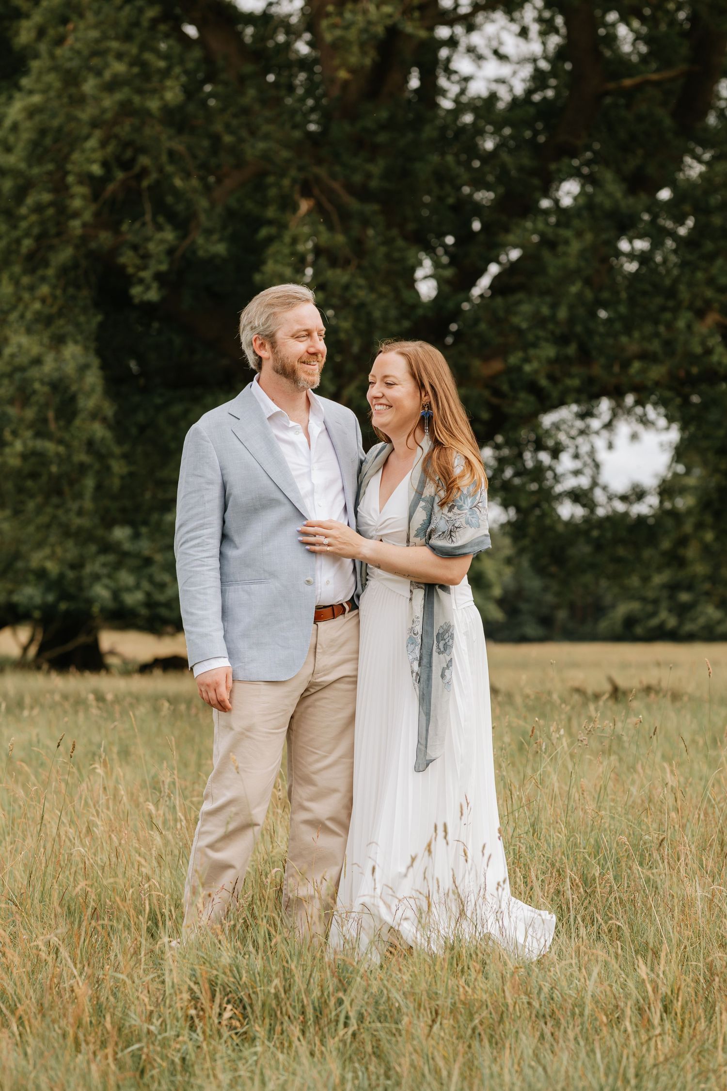 A smiling couple walks through a lush green meadow, the woman in a white dress and man in a light blue blazer.