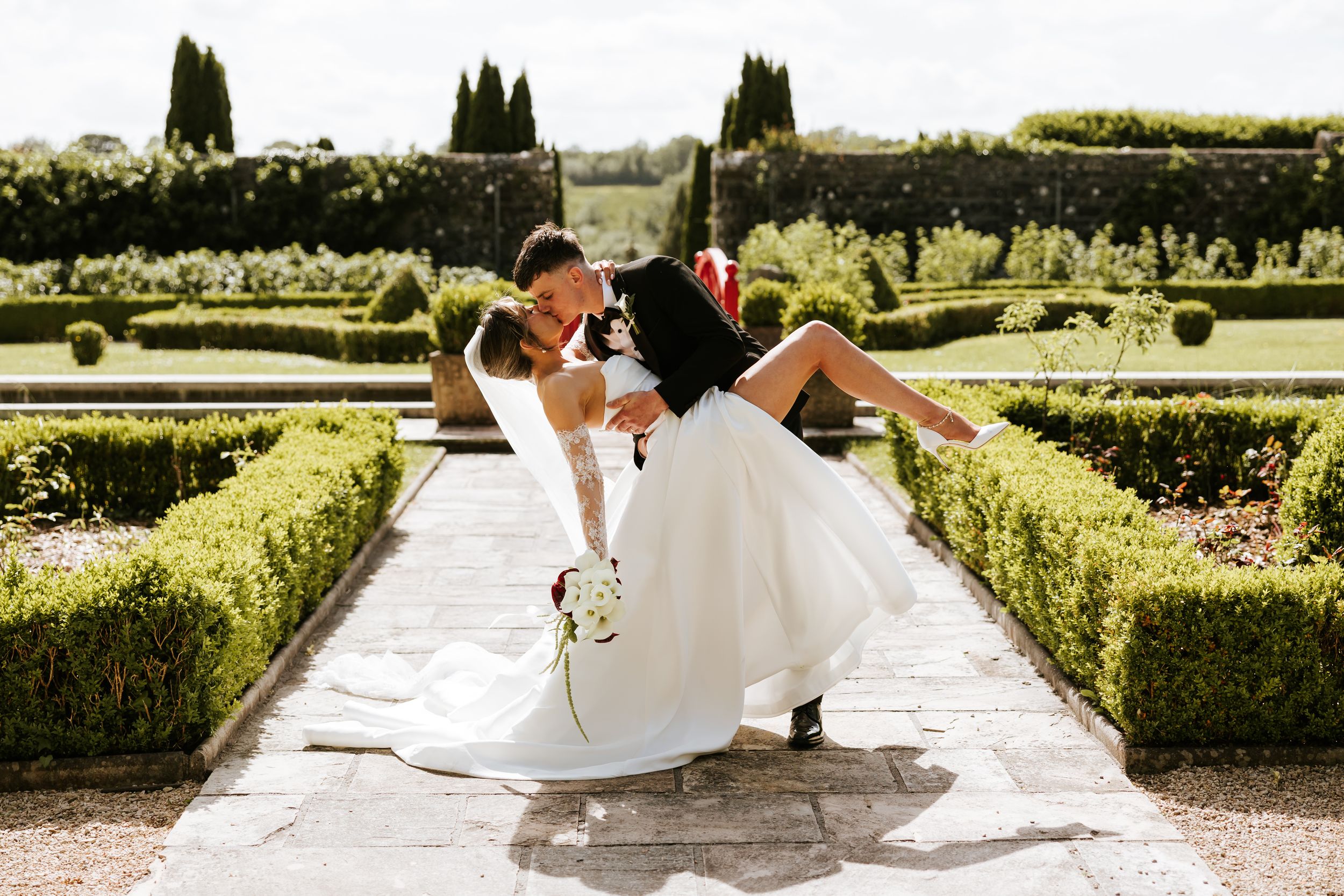 Groom dips bride in elegant formal garden, white wedding dress, lush hedgerows, romantic outdoor ceremony photo.