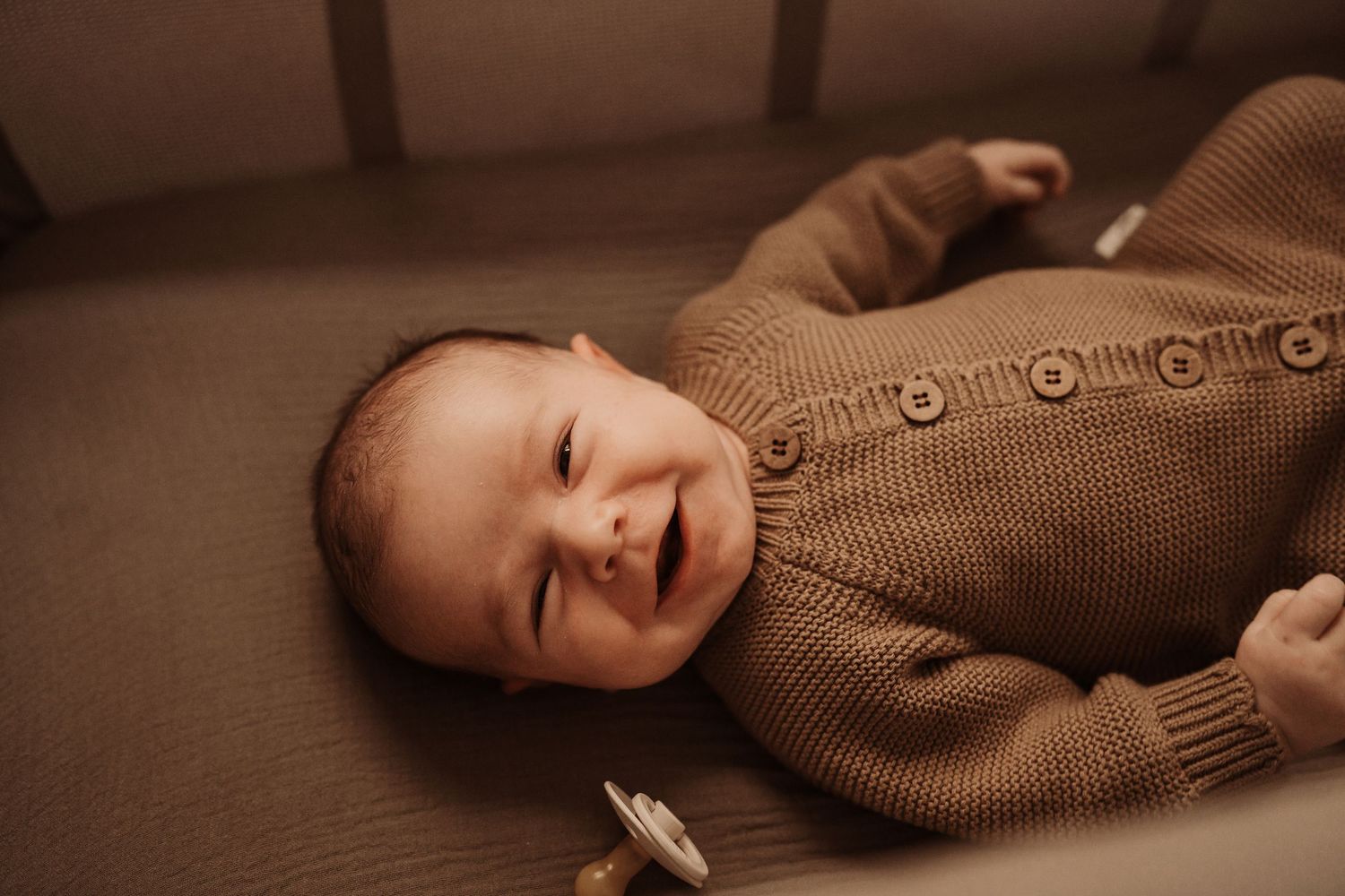 Smiling baby in a brown knit romper lying on a soft surface in a cozy crib setting during a lifestyle newborn session in Chicago