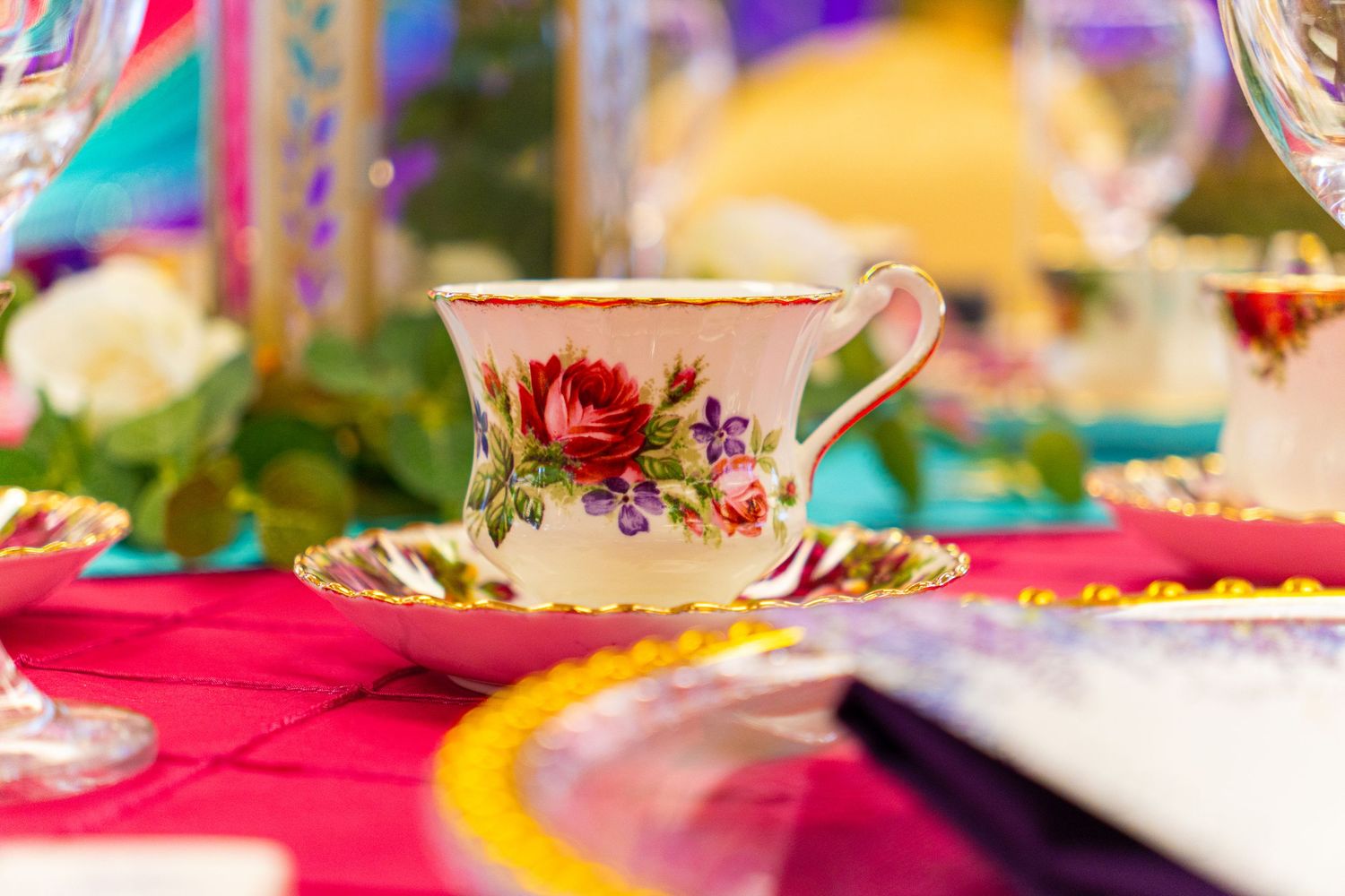 Elegant floral teacup and saucer with gold trim on a colorful table setting with wine glasses and flowers.