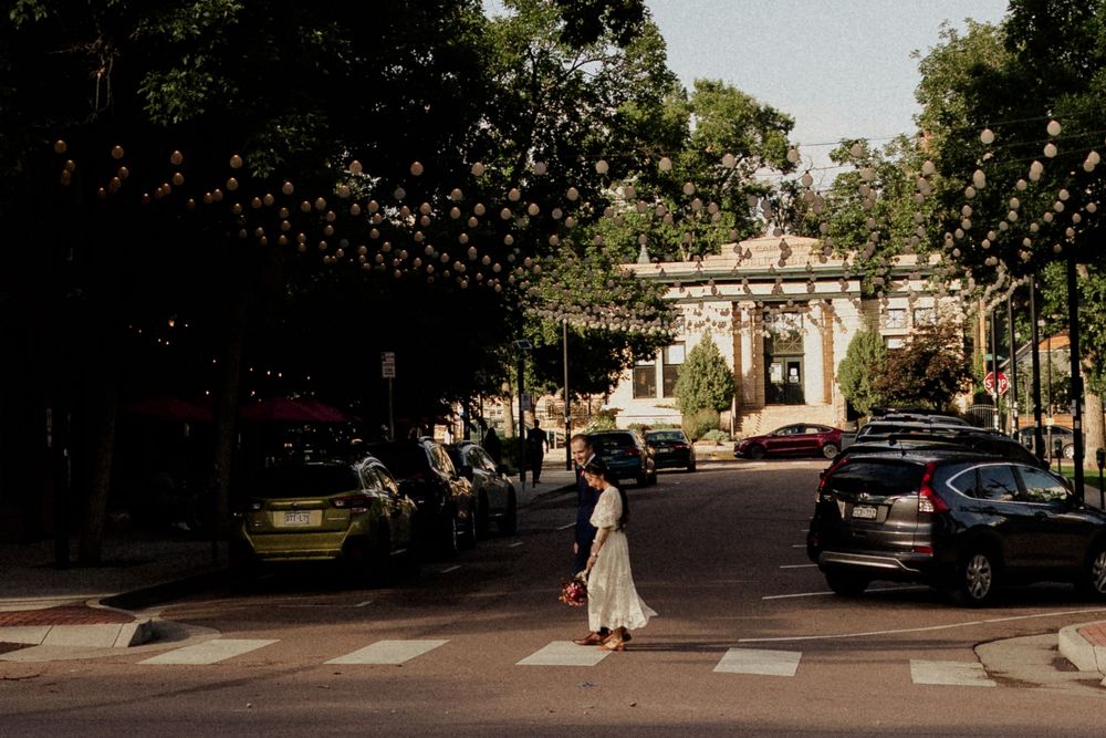Woman crossing a zebra crossing on a tree-lined street with parked cars and decorative string lights overhead.