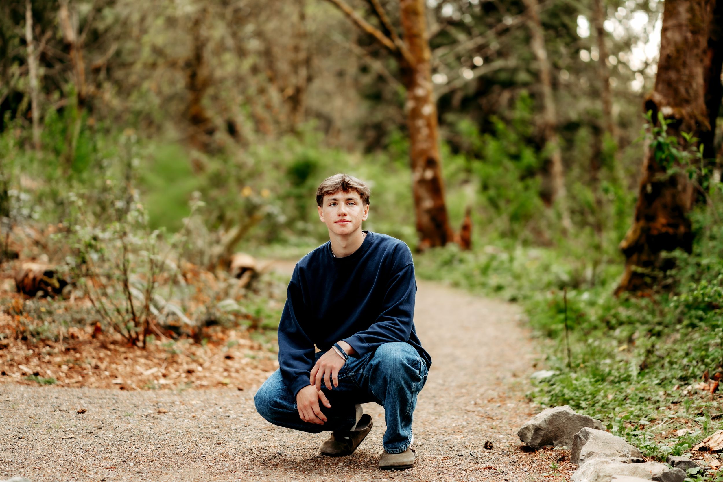 Young man crouching on a forest path beside a small animal, surrounded by lush green trees during senior photos in Puyallup, WA.