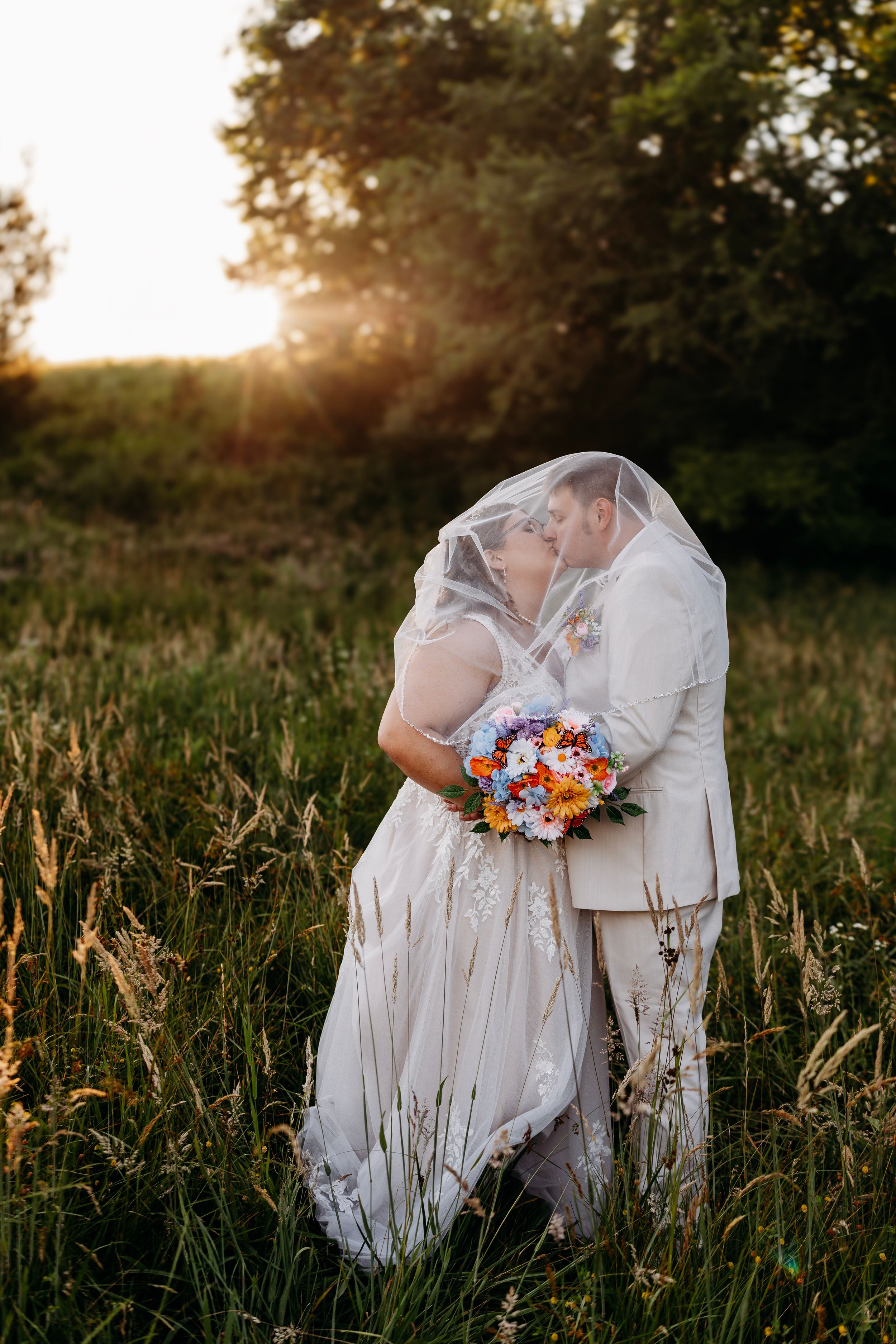Bride and groom sharing a romantic kiss under a veil in a sunlit meadow, bride holding a colorful bouquet at golden hour.