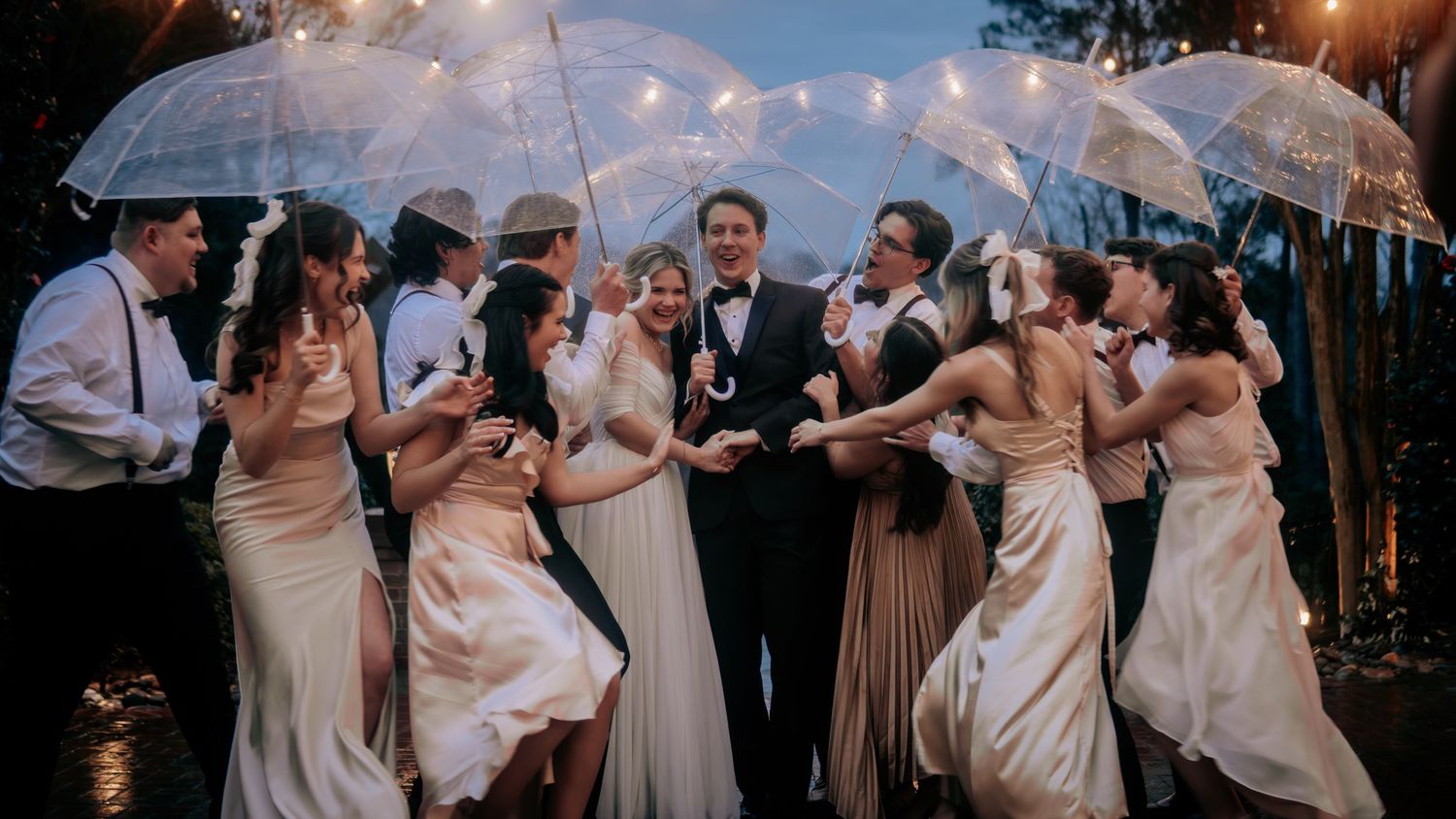 Wedding party celebrates in rain with clear umbrellas, bridesmaids in champagne gowns dancing around bride and groom.