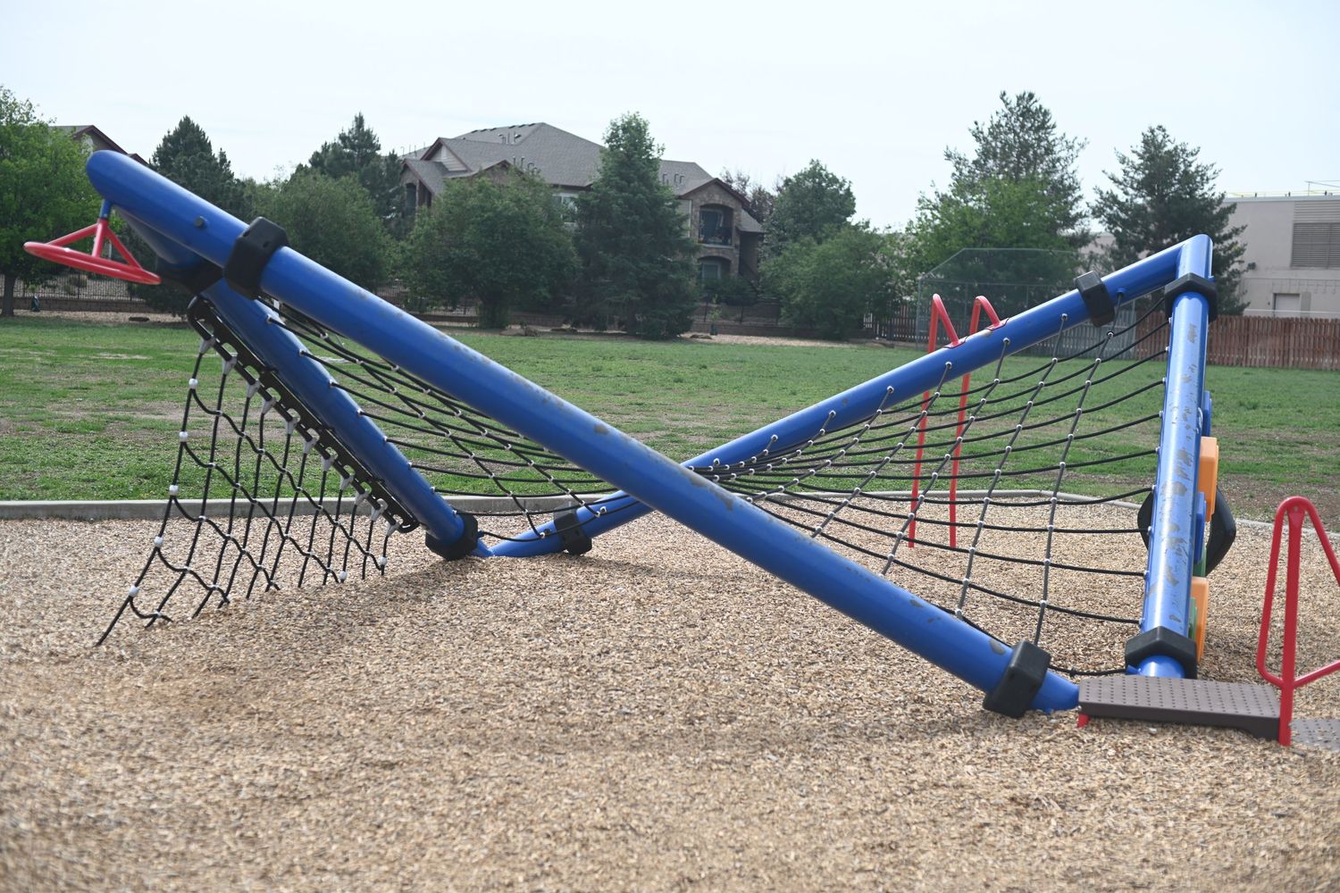Blue playground climbing net structure with rope ladders on sandy ground, surrounded by green grass and residential homes.