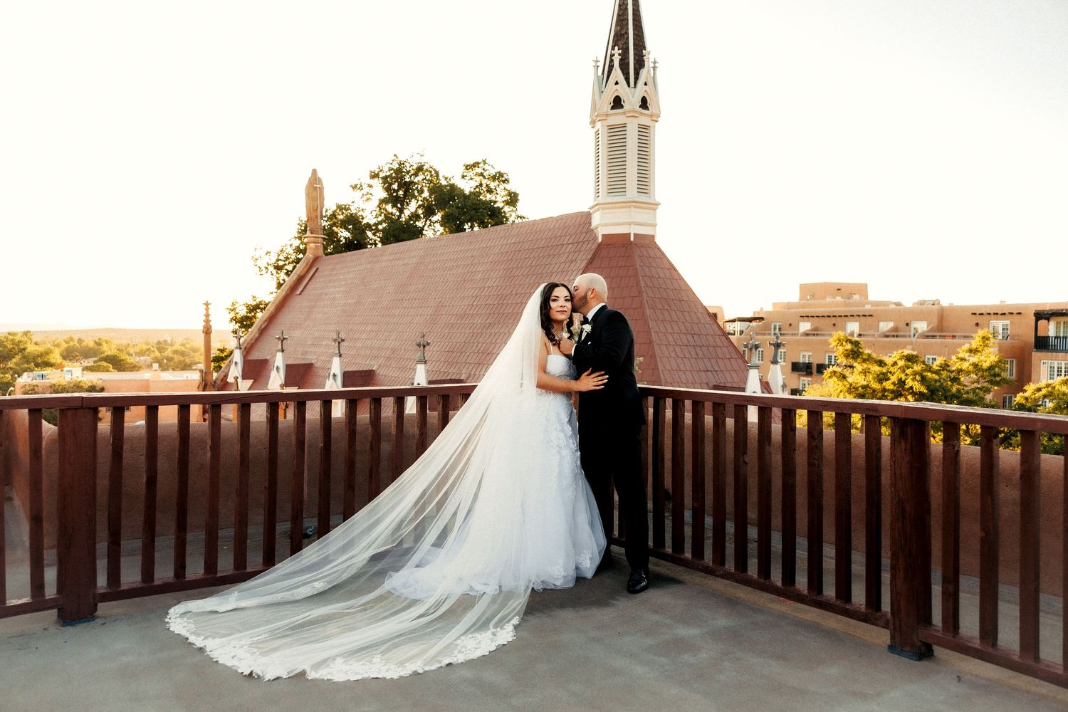Bride and groom embrace on rooftop balcony with flowing cathedral veil, church steeple and cityscape in golden hour background.