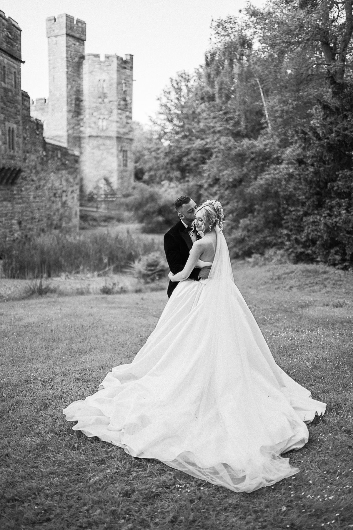 Black and white wedding photo of bride and groom embracing on grass before a historic stone castle.
