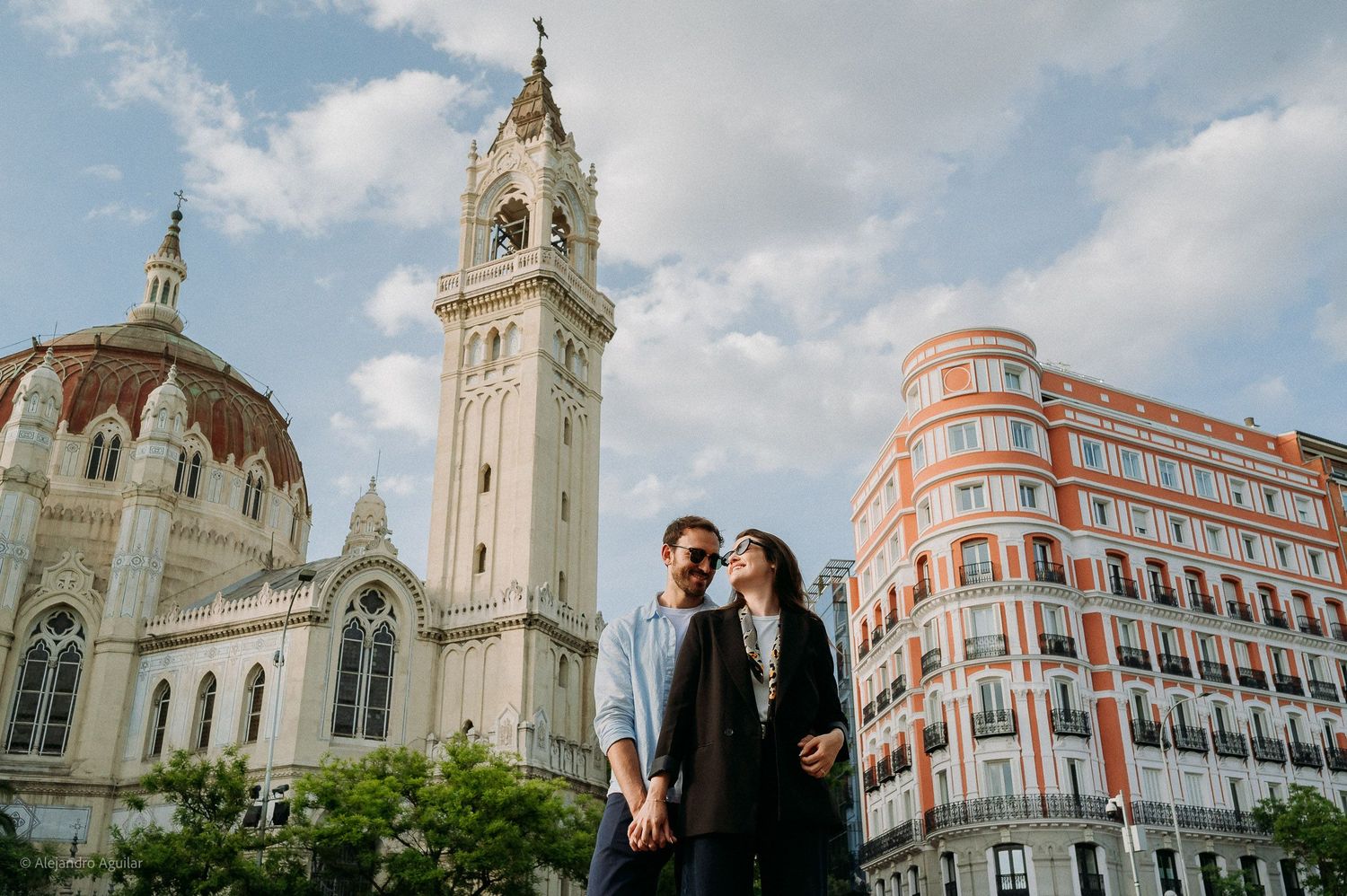Couple smiling together near ornate church with bell tower under blue sky in Madrid.