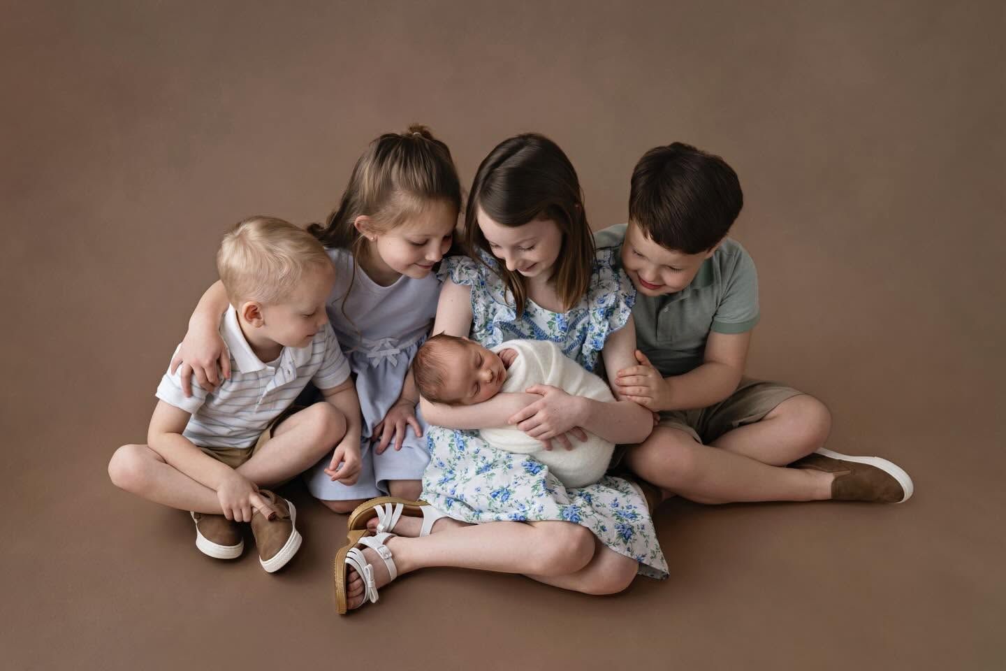 Four siblings sit together on a brown backdrop, lovingly gathered around a newborn baby in a white wrap.