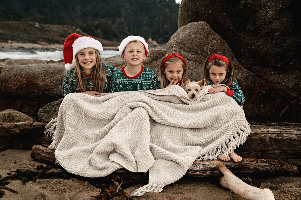 Four children in Christmas sweaters and Santa hats sit on a beach log with a puppy under a cozy white blanket.