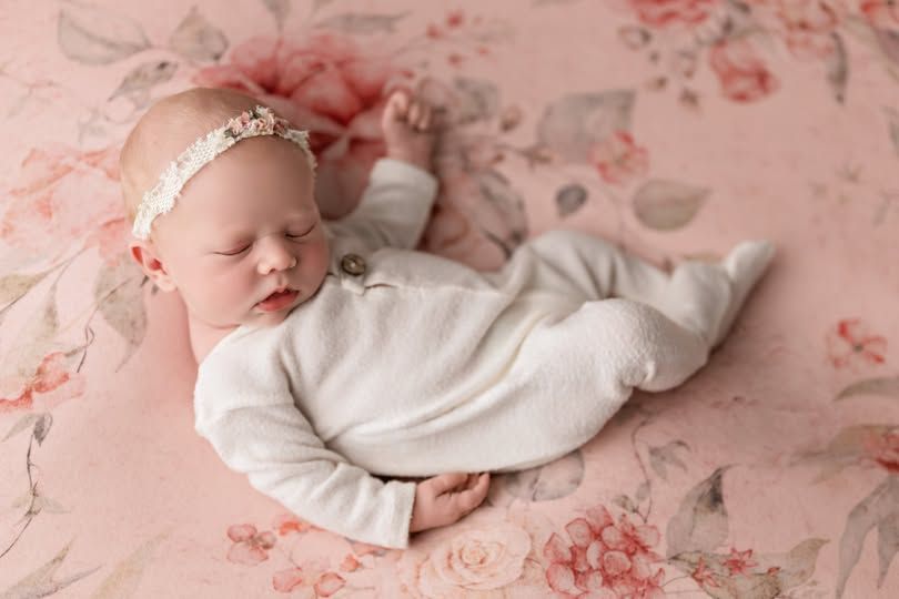 Sleeping newborn baby in white outfit and floral headband on pink floral blanket.