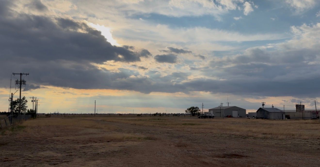 Rural farmstead under dramatic cloudy sky at dusk, with grain silos and sparse flat plains in arid countryside.