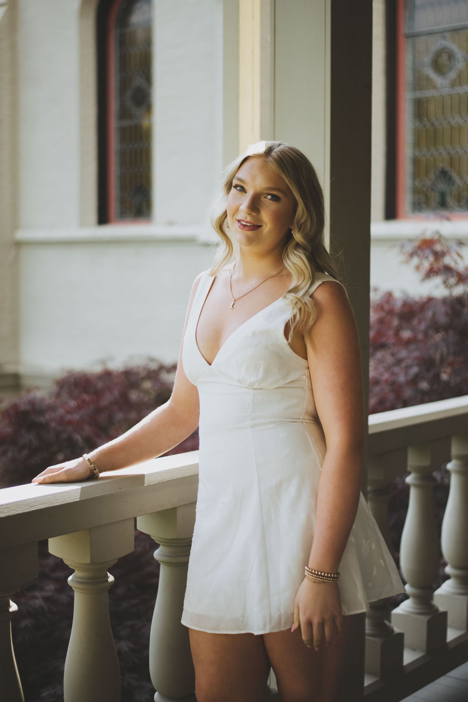 A person in a white summer dress stands on an elegant porch with decorative balustrades and stained glass windows.