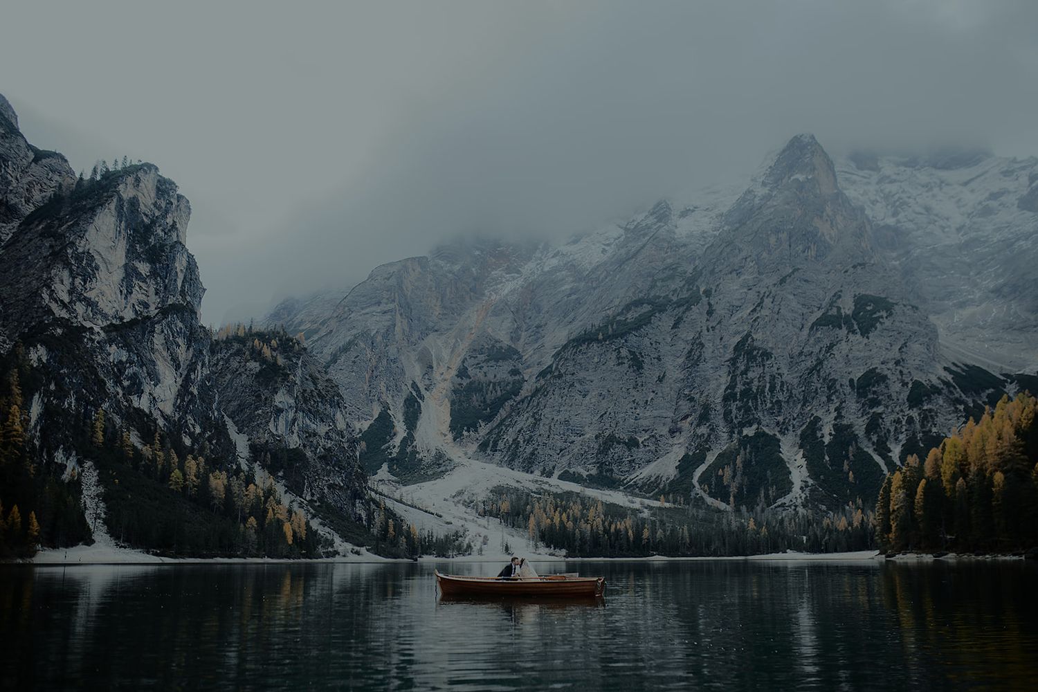 A lone red boat glides across a serene alpine lake surrounded by snow-dusted mountains and autumn trees.