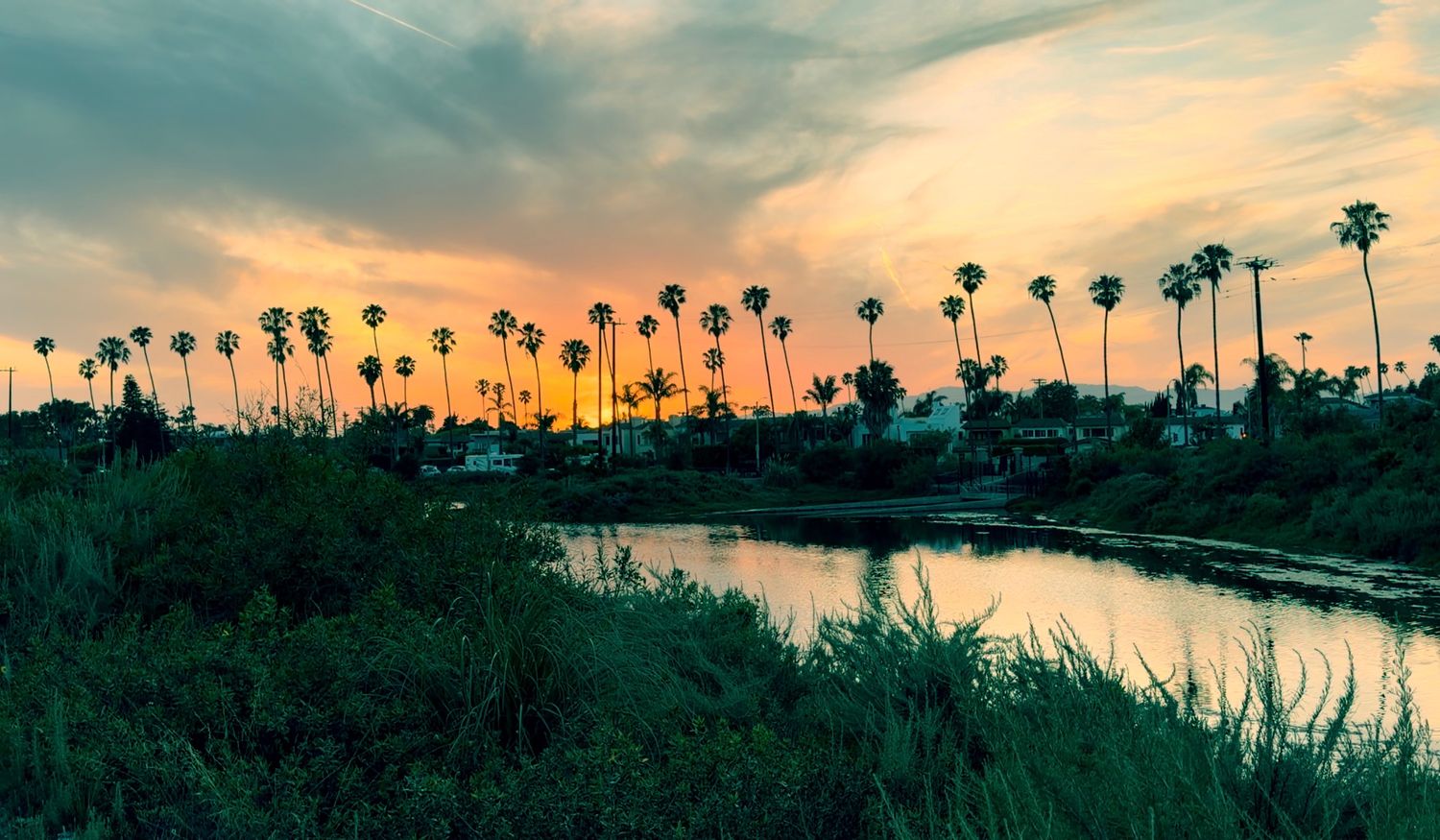 Stunning sunset over a tropical lagoon lined with tall palm trees, reflecting warm orange and pink hues on calm water.