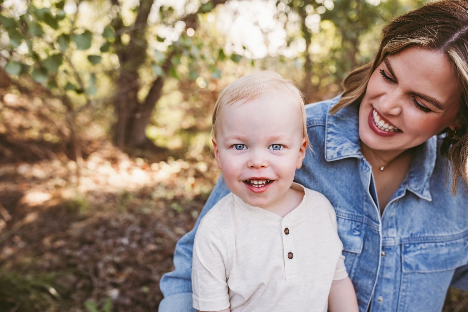 A smiling mother holds her happy blue-eyed toddler boy outdoors in a sunlit forest setting.
