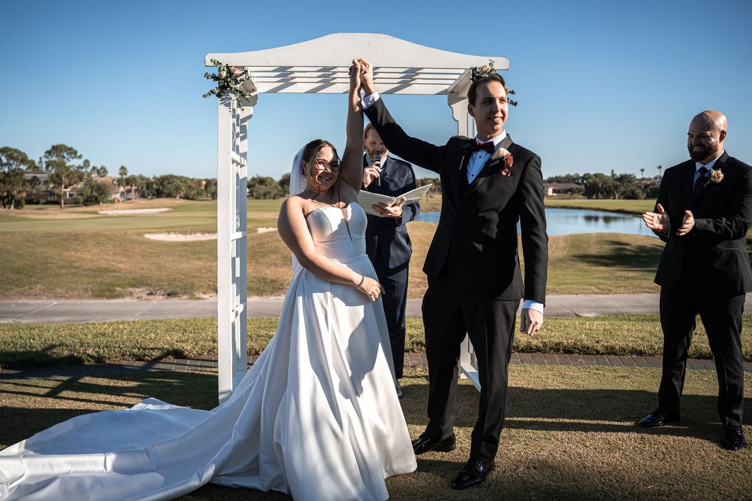 Bride and groom celebrate under white arch at outdoor wedding ceremony on sunny golf course.