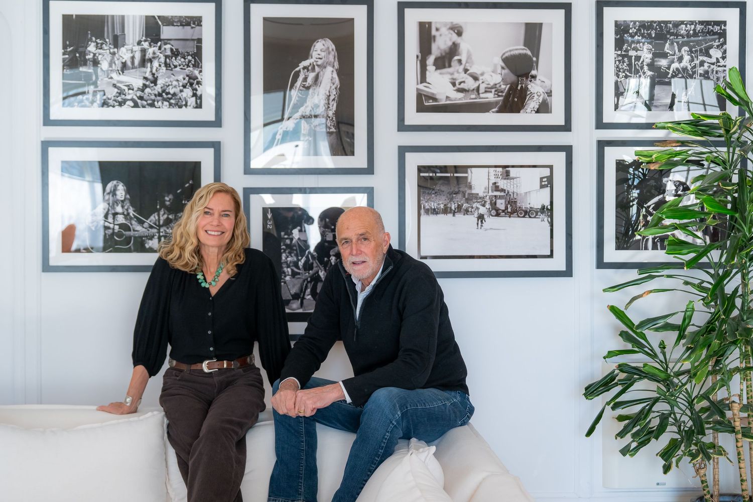 A man and woman sitting together on a white sofa in front of a gallery wall of black and white framed photos.