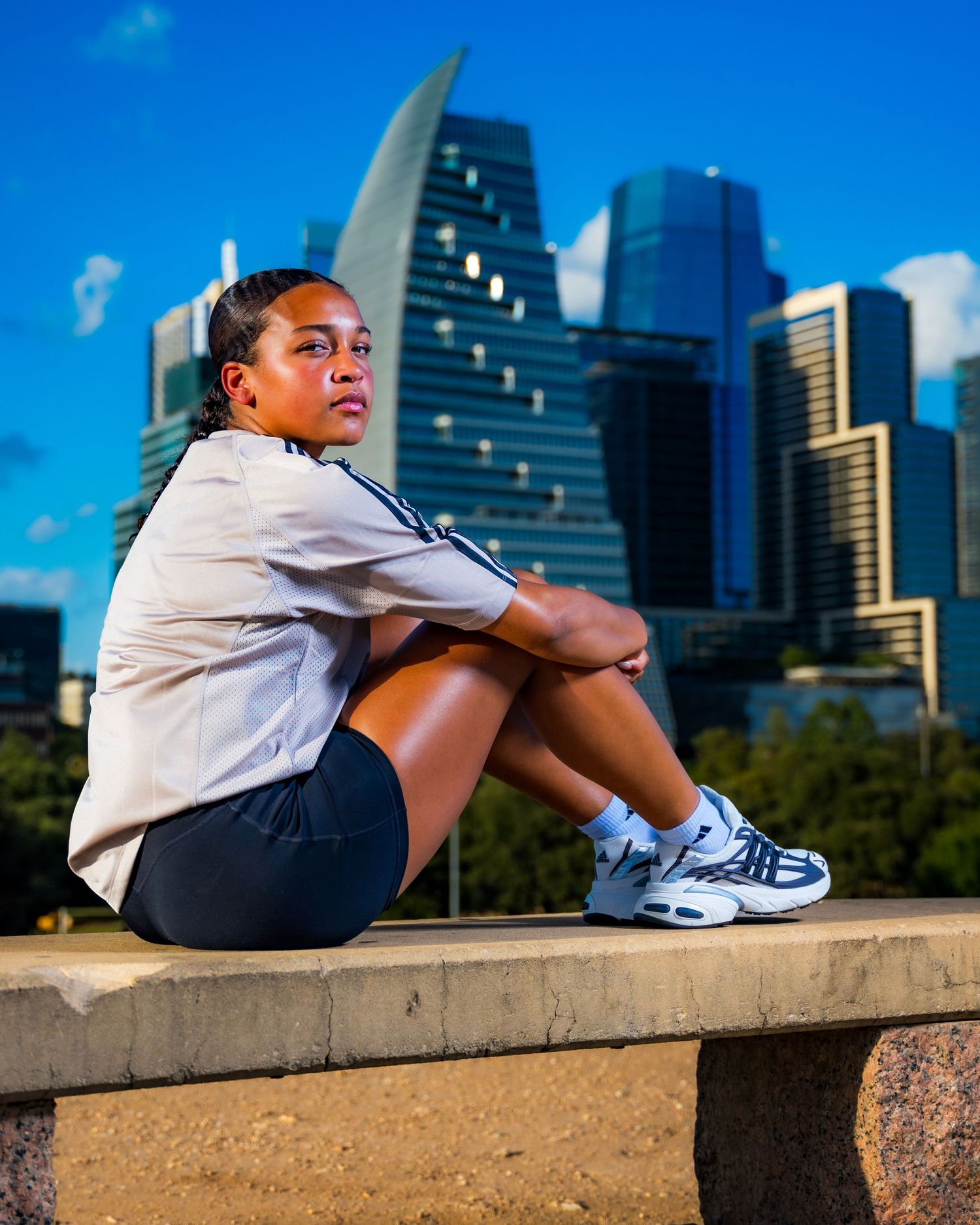 Young woman sitting on stone ledge with Dallas city skyline in background on sunny day.