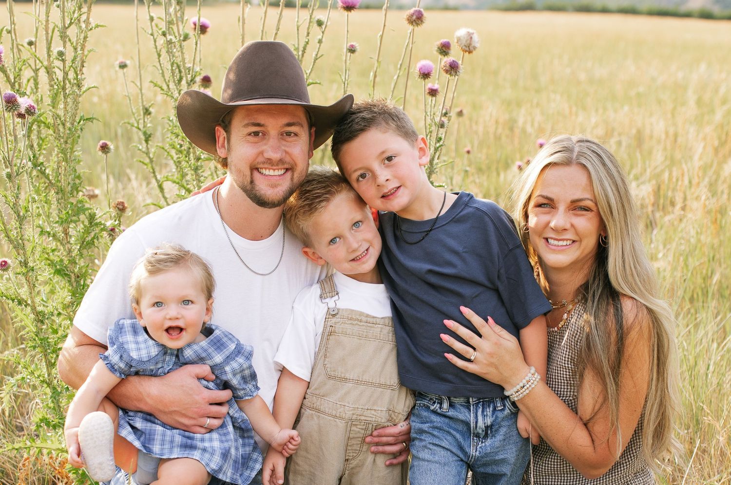 Happy family of five posing outdoors in a wildflower field, parents with three young children smiling for a portrait.