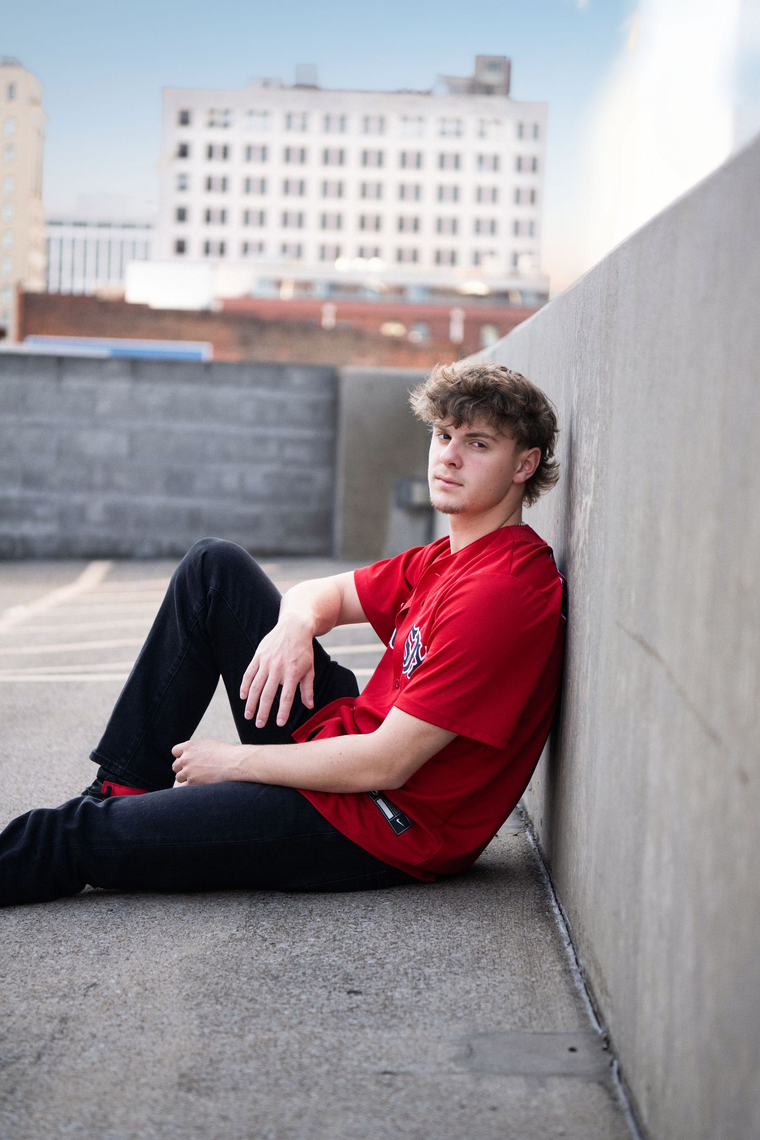 Young man in red shirt sitting against concrete wall on rooftop parking garage with city buildings in background.