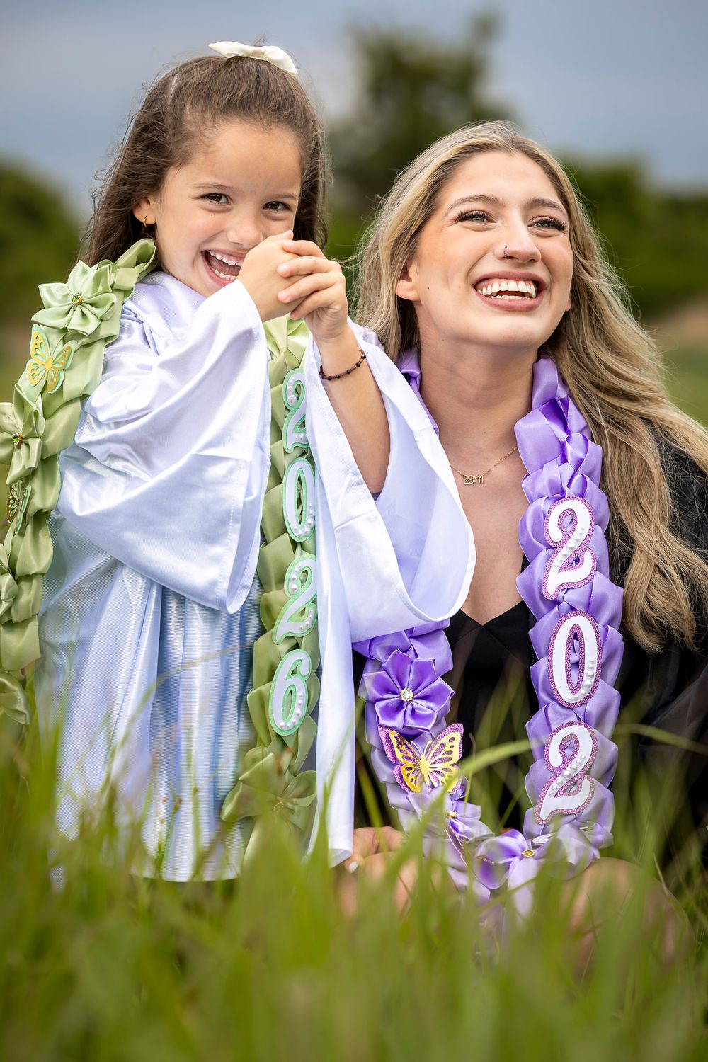 A smiling graduate in black gown with purple 2024 stole poses with a laughing young girl in a green field.
