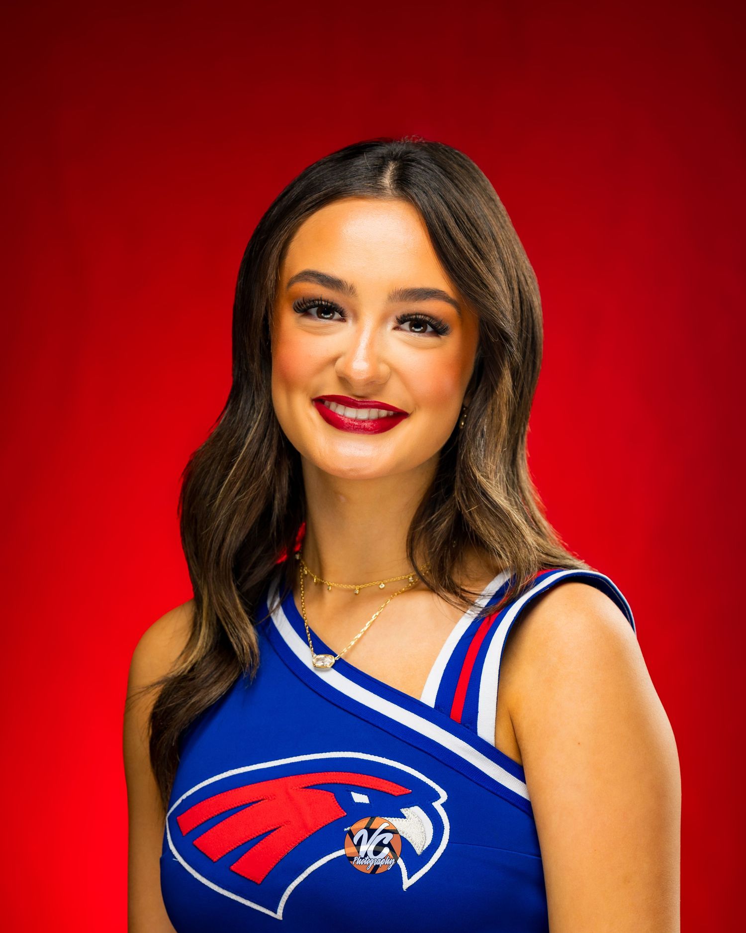 Young woman in blue cheerleader uniform with red hawk logo smiles against vibrant red background.