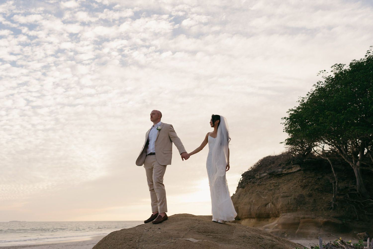 Bride and groom holding hands on a rocky beach at sunset, wearing elegant wedding attire with ocean views.