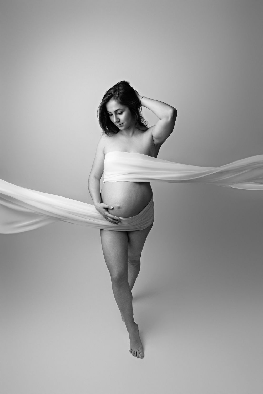 Black and white maternity photo of pregnant woman wrapped in flowing white fabric, posing gracefully in studio.