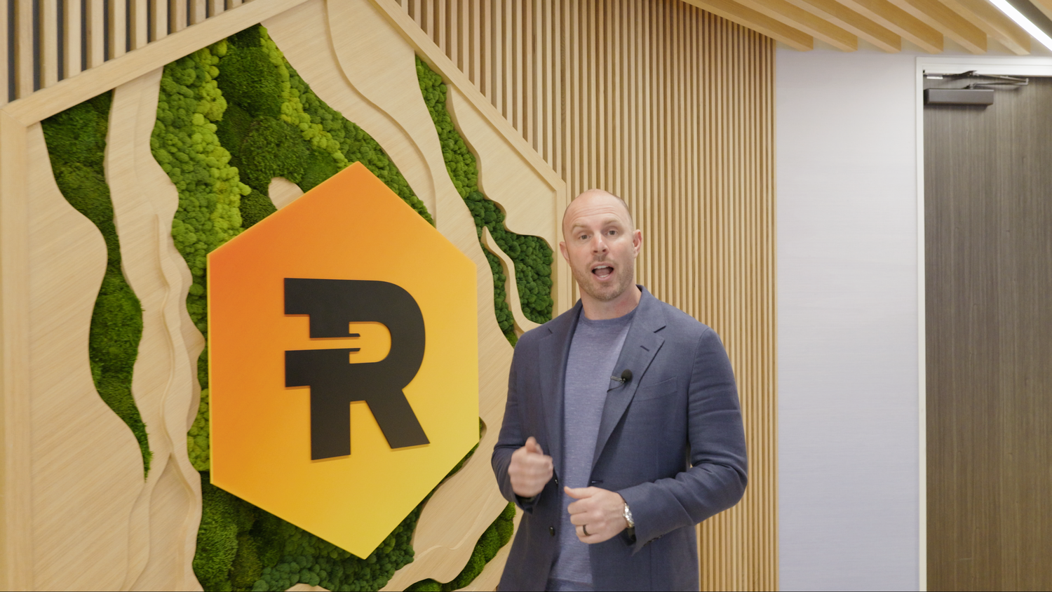 Man in blazer presenting in front of decorative hexagonal logo with green moss wall backdrop.