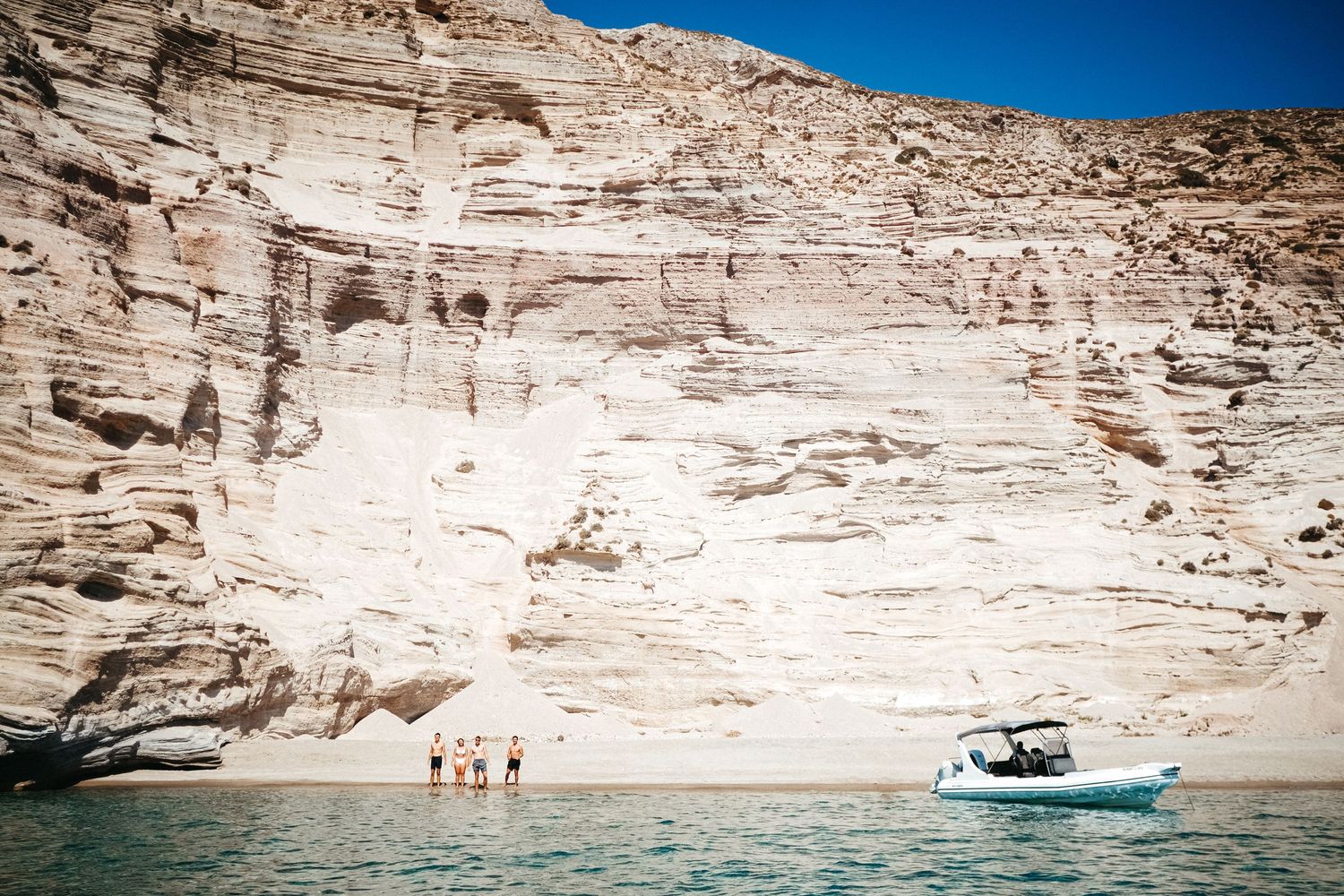 Boat anchored near dramatic white limestone cliffs with swimmers in clear blue water.