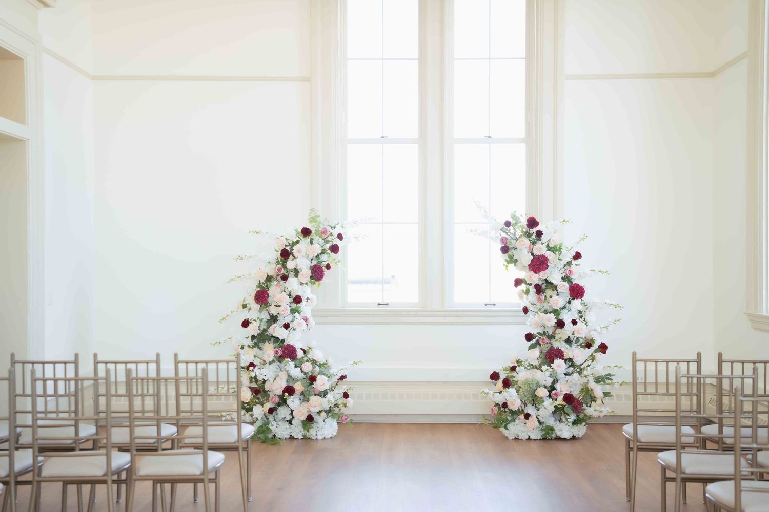 Wedding ceremony setup with floral arrangements framing a window in a bright white room.